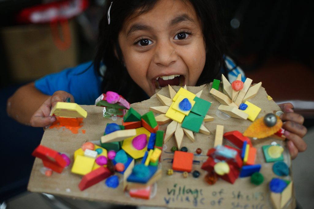 A child smiles excitedly while holding up a colorful cardboard art project covered in painted wooden shapes, beads, and small decorative pieces. The child’s joyful expression and wide open mouth convey pride and enthusiasm for their creation.